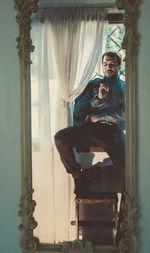 Full length of man sitting on window sill reflecting in mirror