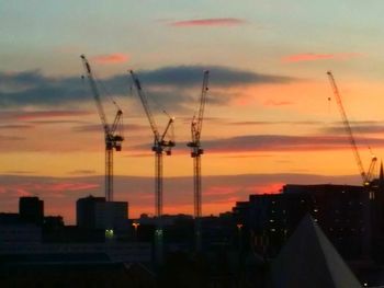 Silhouette cranes and buildings against sky during sunset