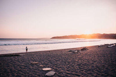 Rear view of woman standing at beach against clear sky during sunset