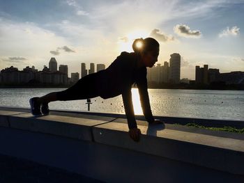 Woman exercising on retaining wall by river in city during sunset