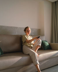 Calm young barefooted lady in casual clothes switching channels with remote controller while resting alone on comfortable sofa in modern apartment in sunlight