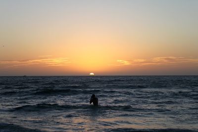 Silhouette man in ocean along edge of beach against sky during sunset