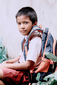 Portrait of cute boy with backpack sitting against wall