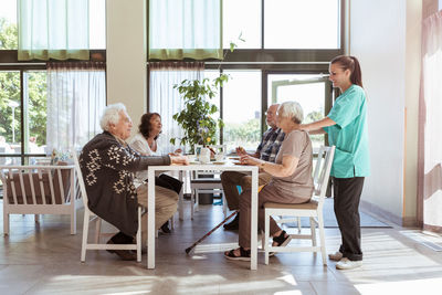 Nurse massaging shoulders of senior woman having breakfast with friends at nursing home