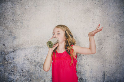 Cute girl playing with tin can phone while standing against wall