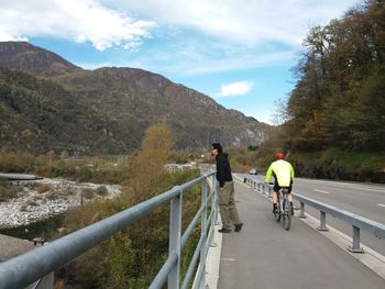 Side view of man standing by cyclist on bridge against mountains