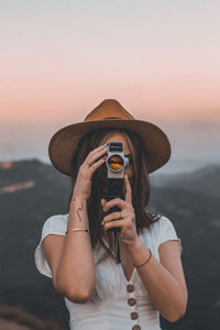 Woman holding camera while standing by sea against sky during sunset