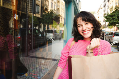 Portrait of smiling young woman holding shopping bag while standing by glass window