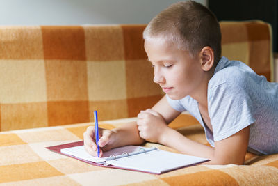 Cute caucasian boy lying on the couch draws on a notebook with a pen or does