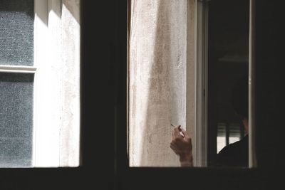 Cropped image of man holding cigarette seen through glass window