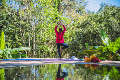 Rear view of woman standing on one leg by water against trees