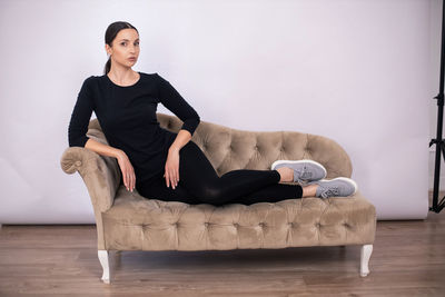 Full length woman poses on a sofa in black tight fitting clothes and sneakers in a studio on a white