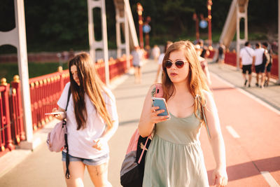 Two young women using smart phones walking on bridge in summer