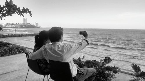 Man with woman taking selfie while sitting on chairs by sea