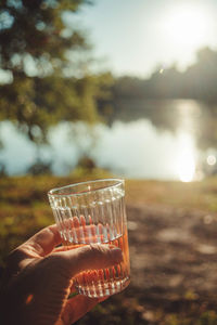 A hand holds a glass of rosé wine near a lake while the sun sets.