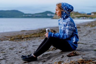 Young woman holding insulated drink container while sitting at beach
