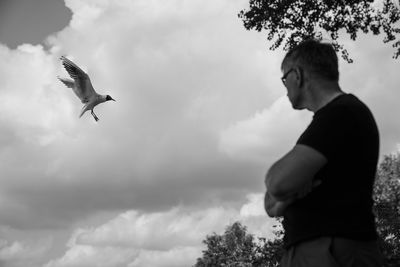 Low angle view of man looking at bird flying against cloudy sky