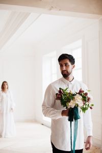 Groom holding flower bouquet with bride standing in background at home
