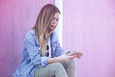 Young woman using phone while standing against pink wall