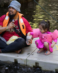 Full length of father and daughter sitting on pier by lake