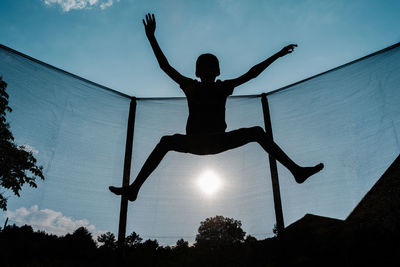 Horizontal front backlighting photo of a silhouette of a barefoot young boy jumping or flying on a trampoline with net and the sun at the background reflecting sunbeam rays on his shadow
