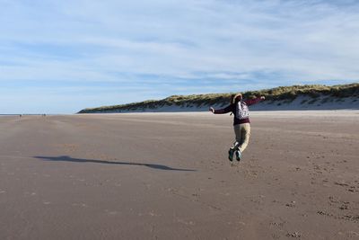 Rear view of woman jumping on beach against cloudy sky