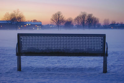 Empty bench in snow against sky during sunset