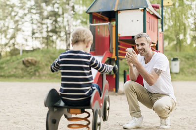 Smiling father photographing son playing on spring ride at playground