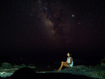 Young woman sitting on rock against sky at night