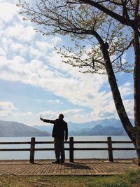 Rear view of man taking selfie while standing on promenade by lake against sky