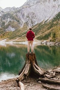 Rear view of man standing on tree stump by lake against mountains