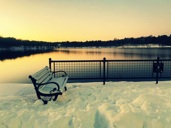 Bench by lake against sky during sunset