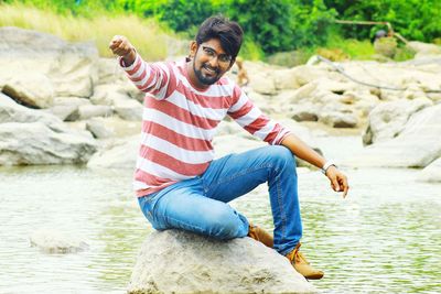 Full length of smiling young man sitting on rock in river