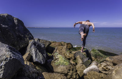 Rear view of shirtless man doing yoga on rock at seashore against clear blue sky