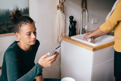 Mid adult woman using mobile phone while boyfriend washing utensils at home