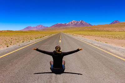 Rear view of carefree woman with arms outstretched sitting on road during sunny day