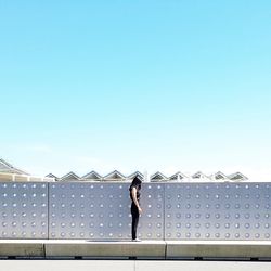 Side view of woman standing by metallic wall against clear blue sky