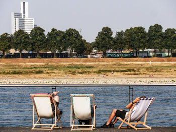 People relaxing on deck chairs at lakeshore against clear sky