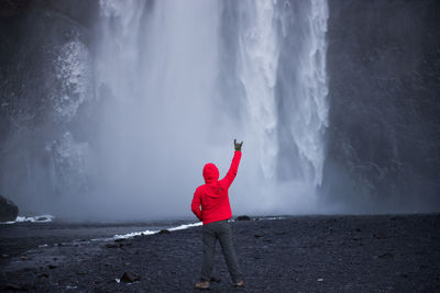 Rear view of man with hand raised looking at waterfall