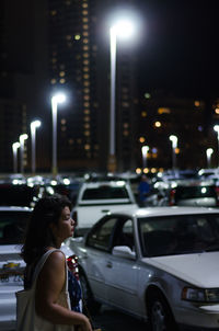 Side view of woman standing at illuminated parking lot during night