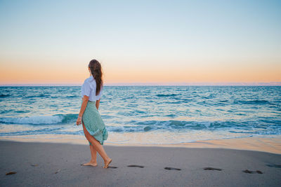 Rear view of woman standing at beach against sky during sunset