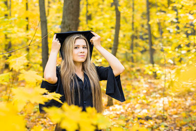 Portrait of young woman wearing graduation gown standing in forest during autumn