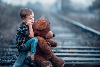 Side view of boy holding teddy bear while sitting on railroad track