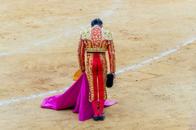 Back view of unrecognizable bullfighter in fancy costume taking off hat after corrida performance while standing on sandy arena