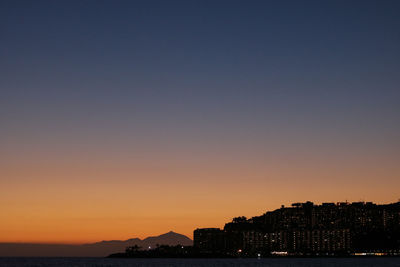 Silhouette buildings by sea against clear sky during sunset