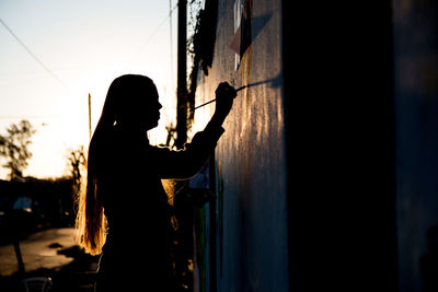 Side view of silhouette woman painting while standing against sky during sunset