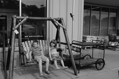 Full length of siblings sitting on swing on footpath