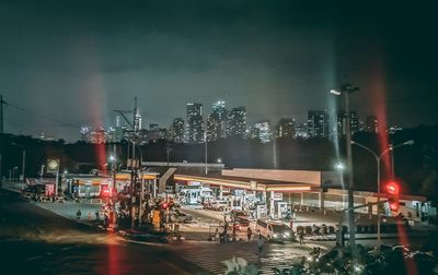 Illuminated street by cityscape against sky at night