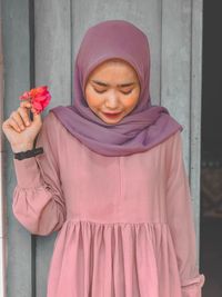 Young woman holding pink flower while standing against wall