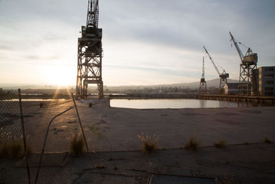 Cranes at harbor against sky during sunset